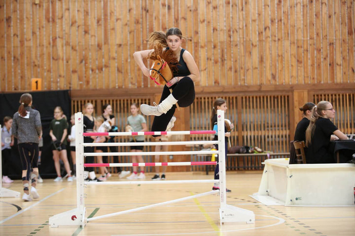 Hobby Horsing competitor jumping a pink and white obstacle in an indoor arena