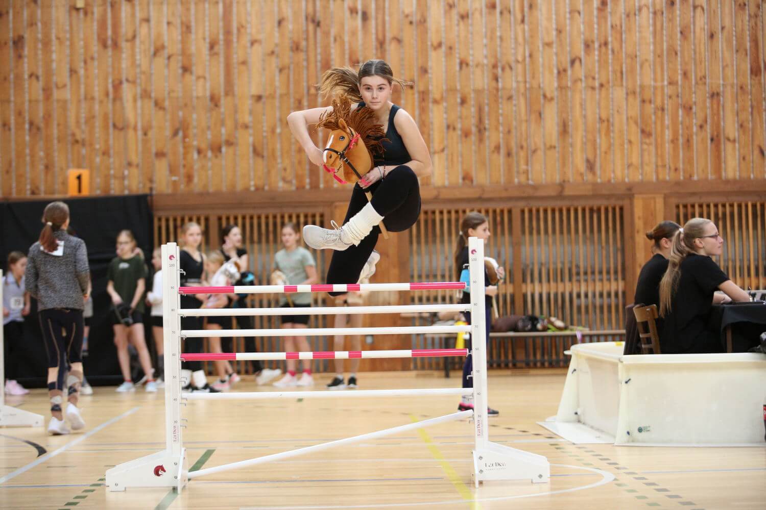 Young participant jumping a pink and white obstacle with a Hobby Horse at an indoor Hobby Horsing event