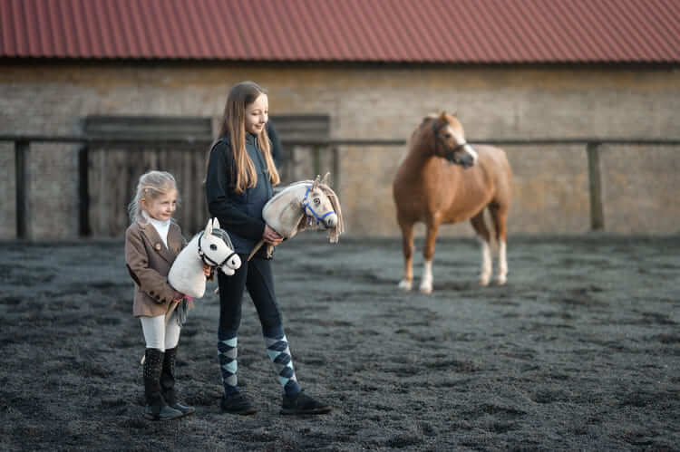 Two children carrying Hobby Horses in an outdoor arena beside a pony