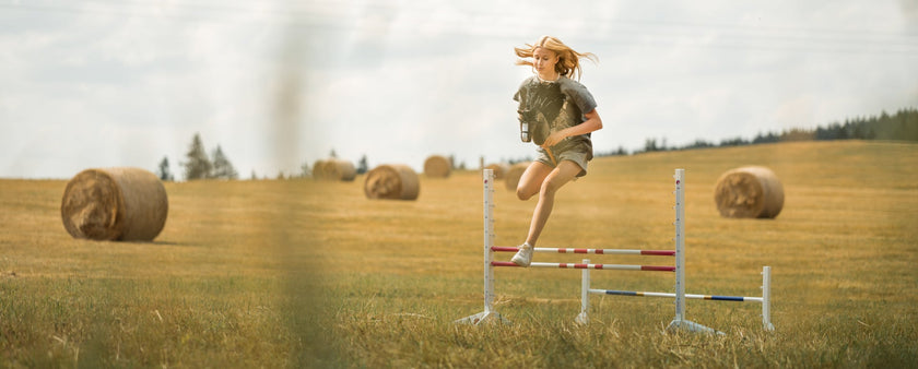 A girl jumps over a hurdle while hobby horsing, holding her hobby horse in a field with round hay bales scattered in the background on a sunny day.