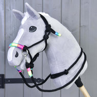 Hobby Horse en peluche gris de Hobby Horse LarDen avec liste blanche et crinière blanche, portant le licou de l’Ensemble de cordes Arc-en-ciel, monté sur un bâton en bois sur fond de panneaux en bois gris.