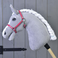 Hobby Horse gris en peluche Hobby Horse LarDen avec crinière blanche tressée, œil noir et Licol framboise (taille M), fixé à un bâton en bois devant un mur à panneaux en bois gris.