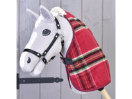 Jouet Hobby Horse gris de Hobby Horse LarDen, portant une couverture d'écurie de Noël à carreaux rouges et une bride noire, monté sur un bâton en bois sur fond de panneaux en bois clair.