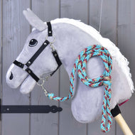Hobby Horse - Corde d'attache tressée turquoise de Hobby Horse LarDen avec tête grise en peluche sur un bâton en bois, bride noire et corde tressée bleu canard, rouge et beige bouclée autour du cou, sur fond en panneaux de bois.