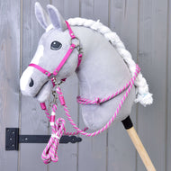 Ensemble Départ Rose (licol + cordelette + longe + rênes) de Hobby Horse LarDen, présenté sur un Hobby Horse en peluche grise à crinière blanche, monté sur un bâton en bois devant un mur en bois gris.