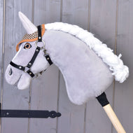 The Hobby Horse LarDen plush hobby horse features a gray head, black bridle, brown plaid "Ear bonnet english brown plaid" for insect protection, white mane, and is mounted on a wooden stick against a wood panel background.