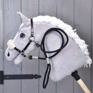 Ensemble Basique de Hobby Horse LarDen, avec Hobby Horse en peluche à crinière blanche, bride noire avec rênes et licou noir sur un bâton en bois, présenté appuyé contre des panneaux en bois gris.