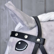 Gros plan sur la tête d’un cheval gris en peluche portant la bride complète noire avec diamants de Hobby Horse LarDen. Le cheval a une crinière blanche, une oreille dressée, un œil de style cartoon, sur fond de planches en bois clair.