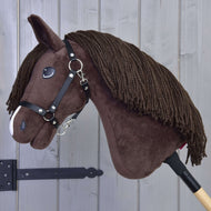 The Hobby Horse LarDen Hobby Horse Chianti with Black Halter, featuring a brown body, yarn mane, and white nose detail, is displayed on a wooden stick against a light gray wooden wall.