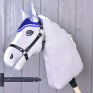 Hobby Horse en peluche de Hobby Horse LarDen avec crinière blanche, bride noire et Bonnet d'Oreilles DeLuxe Bleu pour la protection contre les insectes, monté sur un bâton en bois devant un mur en bois gris clair.