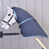 The Hobby Horse LarDen "Blanket with removable neck cover Mandarin" in quilted blue with light blue trim is shown on a gray hobby horse with bridle, set against a wooden plank background.