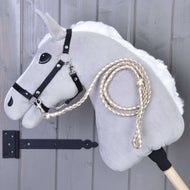 Hobby Horse - Corde d'attache tricotée Double Latte de Hobby Horse LarDen, cheval bâton gris avec bride noire et rênes tressées, présenté devant un mur en bois gris.