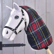 Hobby Horse en peluche au visage blanc et bride noire, portant la couverture d'écurie Édimbourg de Hobby Horse LarDen à carreaux rouges et verts, posé devant un mur en bois gris avec charnière métallique noire visible.