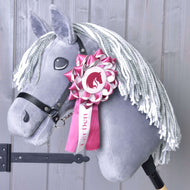 Ruban Hobby Horse LarDen Rose de Hobby Horse LarDen sur un Hobby Horse gris avec crinière blanche en fil, bride noire et ruban de dressage rose et blanc sur la tête, présenté sur fond en bois clair.