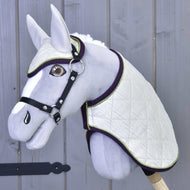 Ensemble Leonardo de Hobby Horse LarDen avec Hobby Horse gris en peluche, couverture blanche matelassée, bonnet d’oreilles assorti, bride noire et rênes sur un bâton en bois, présenté devant un mur en bois gris.