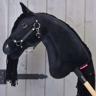 Hobby Horse Maximus avec licol noir de Hobby Horse LarDen, avec yeux brillants en bouton et crinière en fil, monté sur un bâton en bois, présenté devant un mur en planches de bois clair.
