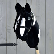 Hobby Horse Merlin avec licol noir de Hobby Horse LarDen, Hobby Horse tacheté noir et blanc inspiré d’un étalon Barock Pinto, avec crinière en fil et bride, monté sur un bâton en bois devant une clôture grise.