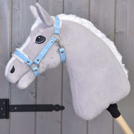 Hobby Horse en peluche grise sur bâton avec crinière blanche portant le licol avec fourrure - similicuir bleu pastel de Hobby Horse LarDen, appuyé contre un mur en bois à panneaux avec charnières métalliques noires.