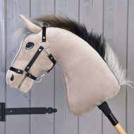 Hobby Horse Piccolino avec licol noir de Hobby Horse LarDen, avec visage beige clair et crinière noire, monté sur un bâton en bois et posé pour le saut d'obstacles devant un mur en bois gris.