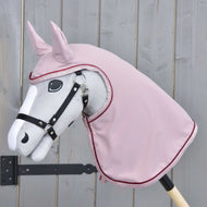 The Hobby Horse LarDen gray hobby horse, mounted on a wooden stick, wears a black bridle and the Raincoat Veloria in pink over its head and neck, posed against a light wooden fence background.