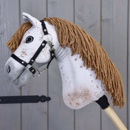 Gros plan du Hobby Horse Rufus avec licol noir de Hobby Horse LarDen, avec visage blanc, crinière brune en fil, licol noir et taches brunes mouchetées, devant un fond en panneaux de bois.