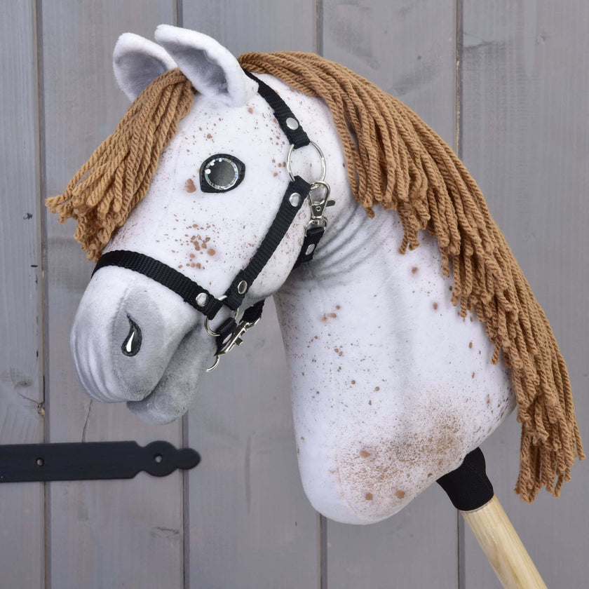Hobby Horse Rufus avec licol noir de Hobby Horse LarDen, avec tête artisanale en tissu blanc moucheté, crinière brune en fil, yeux en bouton et licol noir sur un bâton en bois, présenté devant un fond en bois gris.