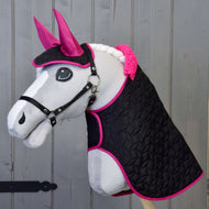 Ensemble Thermo Noir avec Fourrure de Hobby Horse LarDen, Hobby Horse en peluche avec visage blanc, oreilles roses, bride noire et crinière blanche tressée, présenté devant un fond en bois. Il porte une couverture noire matelassée bordée de rose vif.