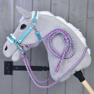 Ensemble Combinaison Parfaite Turquoise de Hobby Horse LarDen sur un Hobby Horse gris en peluche avec crinière blanche, bride turquoise, licol rembourré de fourrure et rênes en corde roses et bleues, suspendu devant un mur en bois gris.
