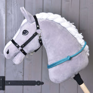 Cordéo Turquoise de Hobby Horse LarDen, Hobby Horse gris en peluche avec crinière blanche en fil, bridon noir, rênes en corde bleue et cordéo turquoise fait main pour monter sans bride, présenté sur un bâton devant un mur en bois.