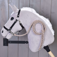 Hobby Horse - Corde d'attache tricotée Amande de Hobby Horse LarDen, cheval en peluche gris clair avec crinière blanche, bride noire et longe amande tricotée à la main, présenté sur un bâton en bois devant un mur en bois gris.