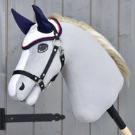 The Hobby Horse LarDen handmade grey Hobby Horse features Ear Bonnet Number 1—white with navy ears for insect protection—a white mane, black bridle, and is mounted on a wooden stick against a light wood panel background.