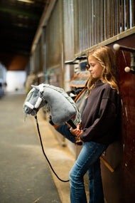 Une fille en sweat marron foncé s’appuie contre les boxes dans une allée d’écurie en tenant le Hobby Horse Calypso avec Licol noir de Hobby Horse LarDen, tête en peluche grise et crinière en fil gris pâle, avec le licol Teddy Classic doublé de fourrure et la longe noire