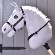 The Cordeo black by Hobby Horse LarDen is a gray plush hobby horse with a white mane on a wooden stick, featuring a black bridle, reins, and handmade cordeo, shown against a gray wooden wall background.