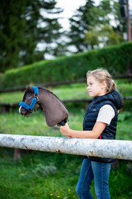 Une fille en gilet matelassé bleu marine se tient derrière une barrière blanche de paddock en tenant le Hobby Horse Chianti avec Licol noir de Hobby Horse LarDen, une haie verte taillée derrière, tête en peluche marron foncé et crinière en fil foncé, avec le licol Teddy Petrol doublé de fourrure
