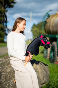 Une fille en pull crème est assise sur un rocher près d’un chemin de prairie en tenant le Hobby Horse Cloony avec Licol noir de Hobby Horse LarDen, tête en peluche noire, étoile blanche et crinière en fil noir, avec le licol noir en similicuir à fourrure rose complète