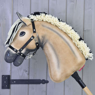 A tan and black Hobby Horse LarDen Colby with a black halter and yarn mane is mounted on a wooden stick against a gray wooden wall.