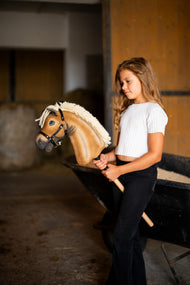 Une fille en haut blanc se tient dans une écurie en tenant le Hobby Horse Colby avec Licol noir de Hobby Horse LarDen, tête en peluche palomino, épaisse crinière en fil crème et licol noir avec ferrures argentées