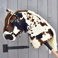 Hobby Horse Dakota avec licol noir de Hobby Horse LarDen, cheval bâton en peluche avec taches marron et blanches, bride et crinière en fil inspirées des chevaux mustang sauvages d’Amérique du Nord, présenté devant un mur en bois gris.