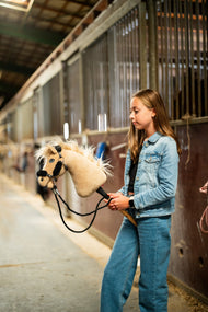 Une fille en veste en jean promène le Hobby Horse Dancing Lady avec Licol noir de Hobby Horse LarDen dans une allée d’écurie, tête en peluche crème, crinière et toupet en fil pâle, avec le licol noir en similicuir à fourrure noire complète et les rênes noires