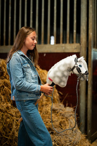 Une fille en veste en jean se tient devant des balles de paille dans une écurie en tenant le Hobby Horse Elektra avec Licol noir de Hobby Horse LarDen, tête en peluche blanche pommelée, œil bleu et crinière en fil blanc, avec le licol Graphite Full en similicuir avec fourrure et la longe tricotée