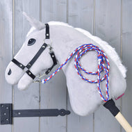Hobby Horse - drapeau à rayures corde CZ de Hobby Horse LarDen sur un Hobby Horse au corps gris, crinière blanche, bride noire et laisse tressée rouge, blanche et bleue, monté sur un bâton en bois et appuyé contre un mur en bois gris clair.