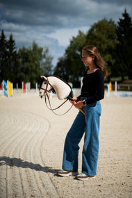 Une fille en haut noir se tient dans une carrière de sable sous un ciel orageux en tenant le Hobby Horse Frederik LD avec Licol noir de Hobby Horse LarDen, des barres d’obstacle colorées derrière, tête en peluche crème et crinière en fil marron foncé, avec la bride marron complète avec muserolle