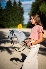 Une fille en haut rose promène le Hobby Horse Grace avec Licol noir de Hobby Horse LarDen dans une carrière de sable, tête en peluche grise et crinière en fil gris pâle, avec la bride De Luxe complète à deux frontaux