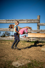 Une fille en tenue d’équitation traverse un paddock sablonneux avec le Hobby Horse LarDen Pinkie avec licol pailleté de Hobby Horse LarDen, tête en peluche rose à crinière en fil rose et blanc, présenté avec le Licol turquoise