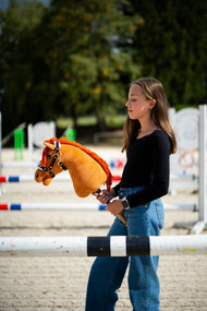 Une fille en haut noir se tient derrière une barre d’obstacle dans une carrière de sable en tenant le Hobby Horse Little Angel avec Licol noir de Hobby Horse LarDen, tête en peluche alezane et crinière en fil rouge vif, avec le licol Espresso en similicuir avec fourrure
