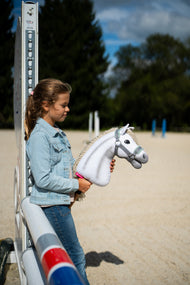 Une fille en veste en jean se tient près d’un chandelier d’obstacle dans une carrière de sable en tenant le Hobby Horse Little Lady avec Licol noir de Hobby Horse LarDen, tête en peluche blanche et crinière en fil crème, avec le licol gris à fourrure menthe