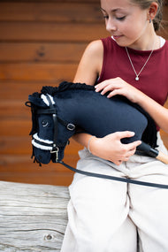 Une fille en haut bordeaux tient dans ses bras le Hobby Horse Mahal avec Licol noir de Hobby Horse LarDen devant un mur en bois, tête en peluche noire, crinière en fil noir et muserolle en fourrure blanche, présenté ici avec la bride complète avec fourrure