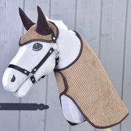 Ensemble Manchester Le Chocolat de Hobby Horse LarDen avec Hobby Horse en peluche au visage blanc, harnais noir, oreilles marron et tissu marron texturé sur la tête et l’encolure, recouvert d’une couverture pour Hobby Horse, monté sur un bâton en bois devant un mur en bois gris.