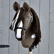 Hobby Horse Monte avec licol noir de Hobby Horse LarDen, fabriqué à la main, avec robe marron et blanche inspirée de l’American Paint Horse, licol noir, longue crinière en fil, monté sur un bâton en bois devant un fond en bois gris.