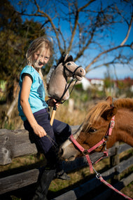 Une fille blonde en haut turquoise est assise sur une clôture de paddock en bois avec le Hobby Horse Piccolino avec Licol noir de Hobby Horse LarDen, tête en peluche beige clair à crinière en fil foncé, un poney alezan derrière elle, présenté avec la Bride complète noire en similicuir
